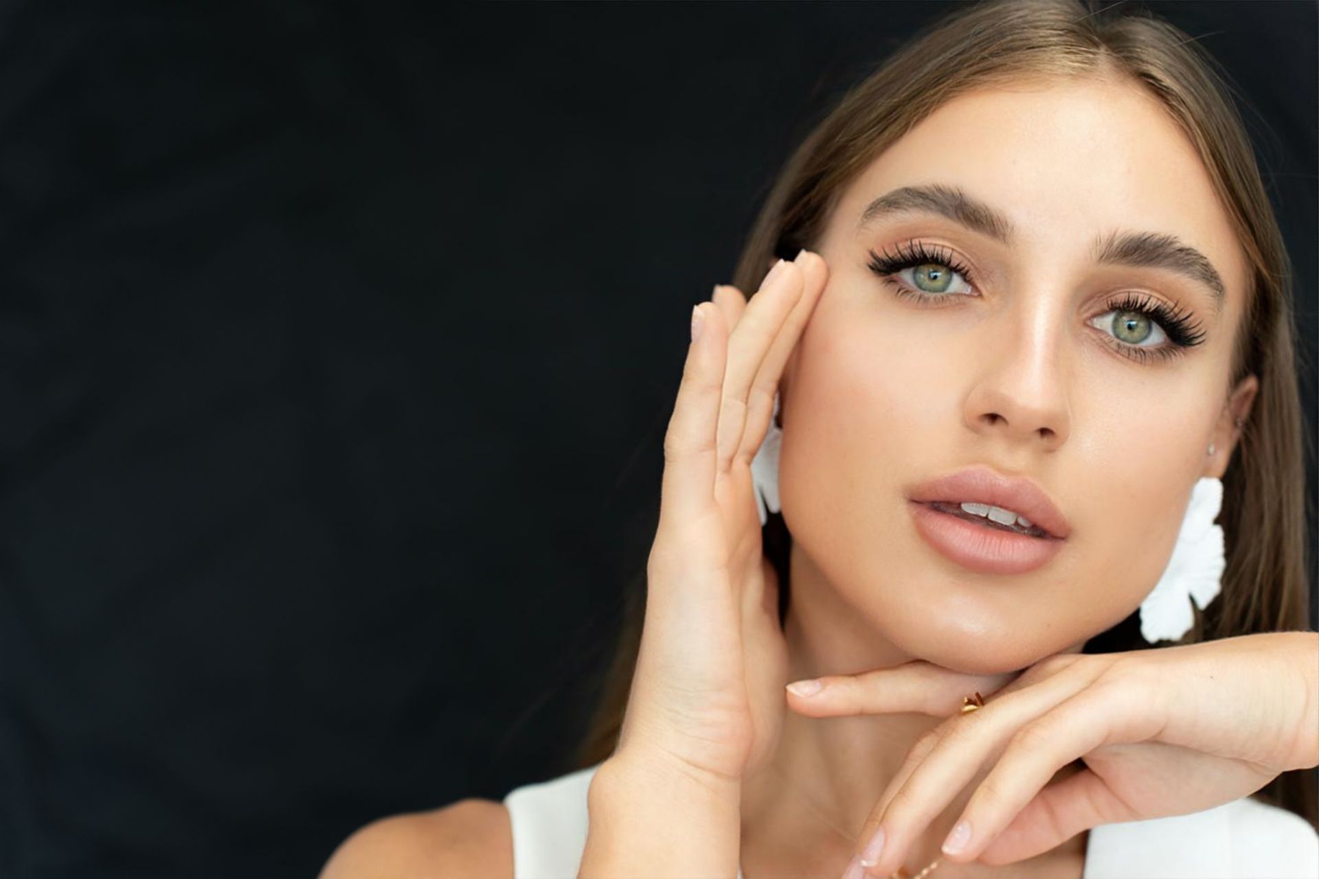 A woman is touching her face with her hands and wearing white earrings.