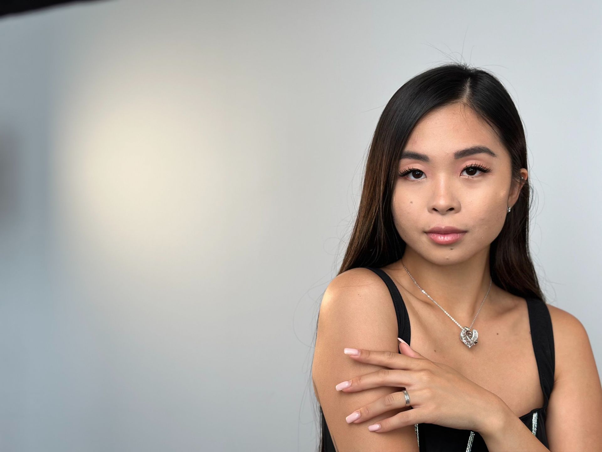 A woman wearing a necklace and a ring is standing in front of a white wall.