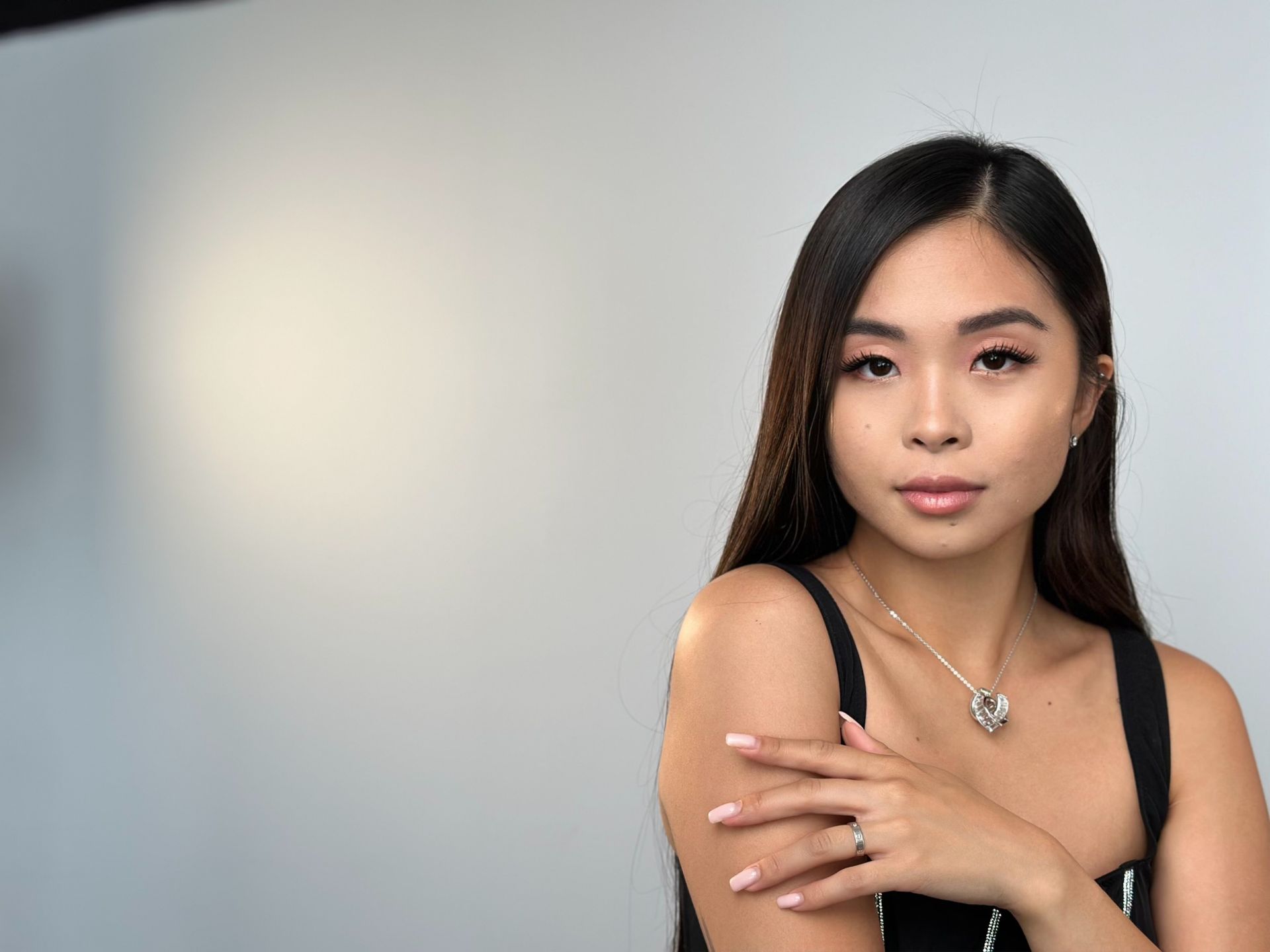 A woman wearing a necklace and a ring is standing in front of a white wall.