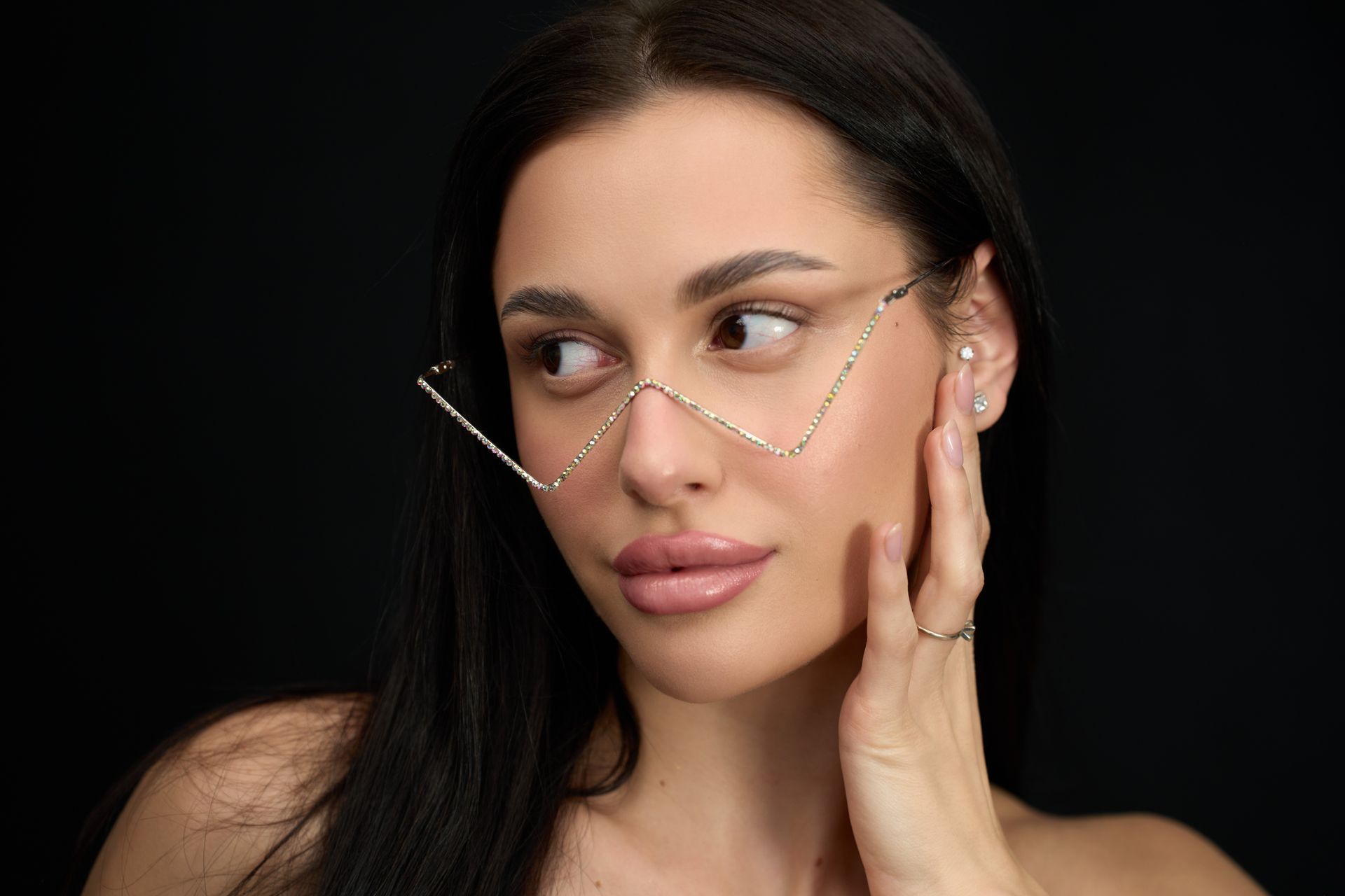 Woman with dark hair wearing gold geometric eyeglasses, looking to the side with hand on face, against black background.
