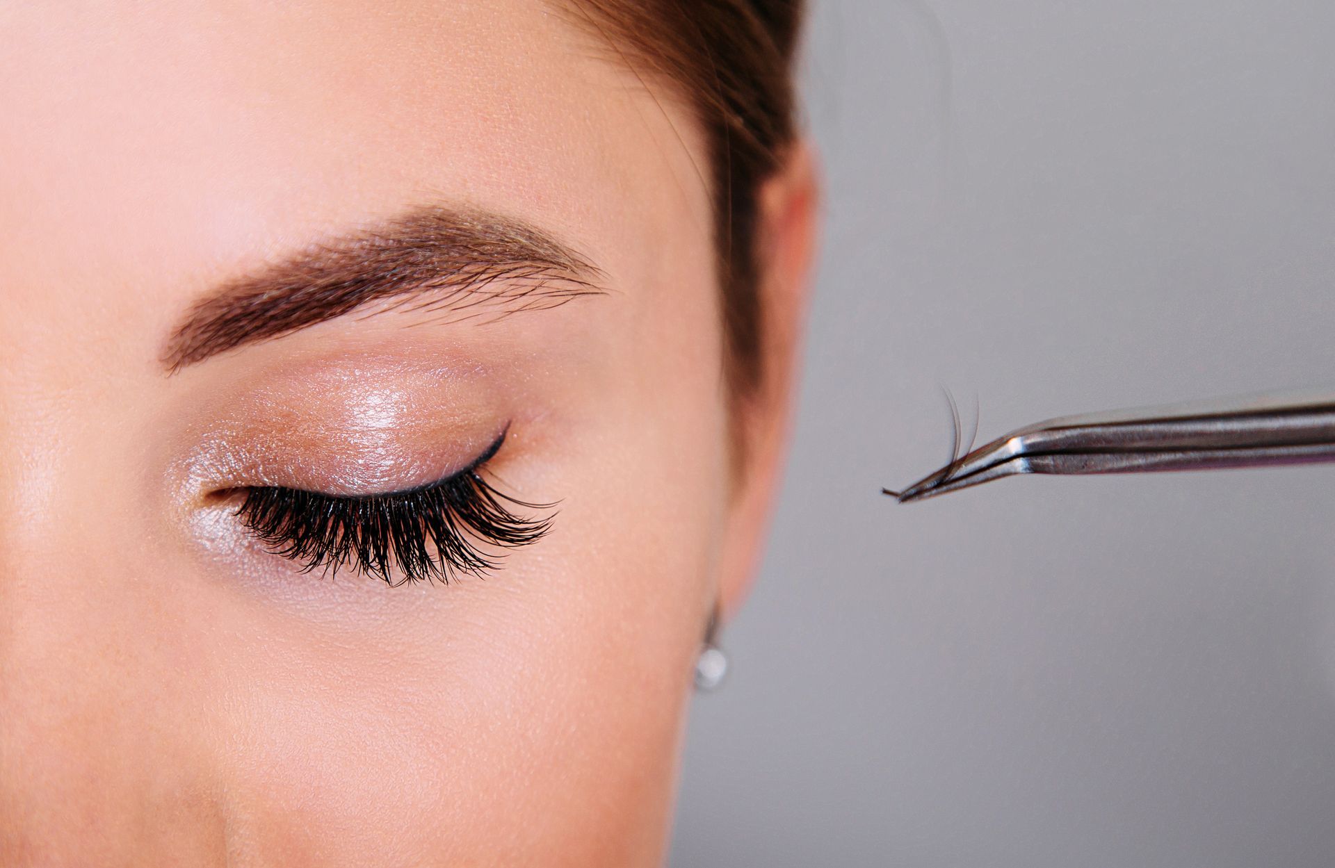 Eye with long, black lashes; tweezers holding a small lash extension; pale skin, neutral background.