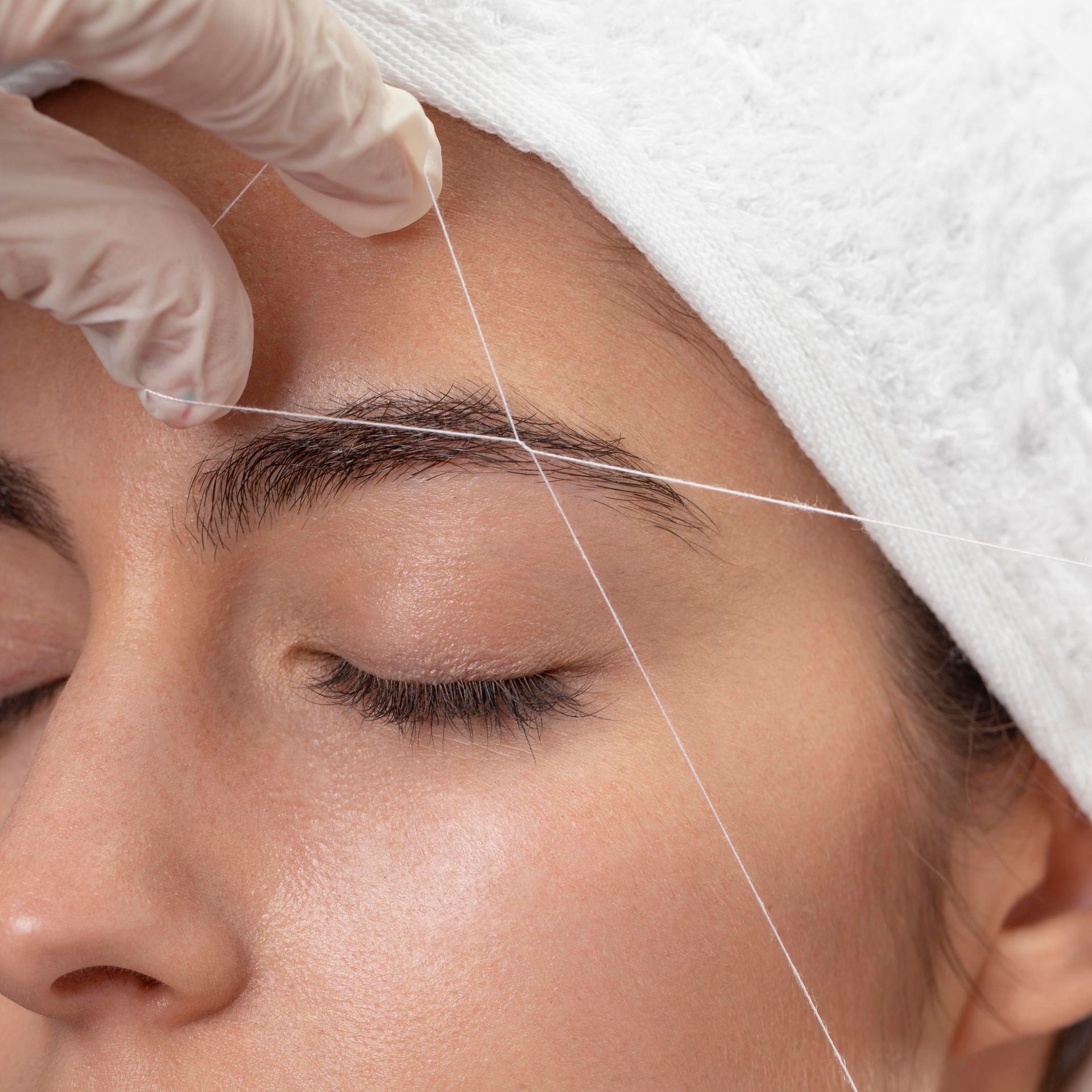 A hand in a white glove uses a thin white thread to shape and remove hair from a person's eyebrow.