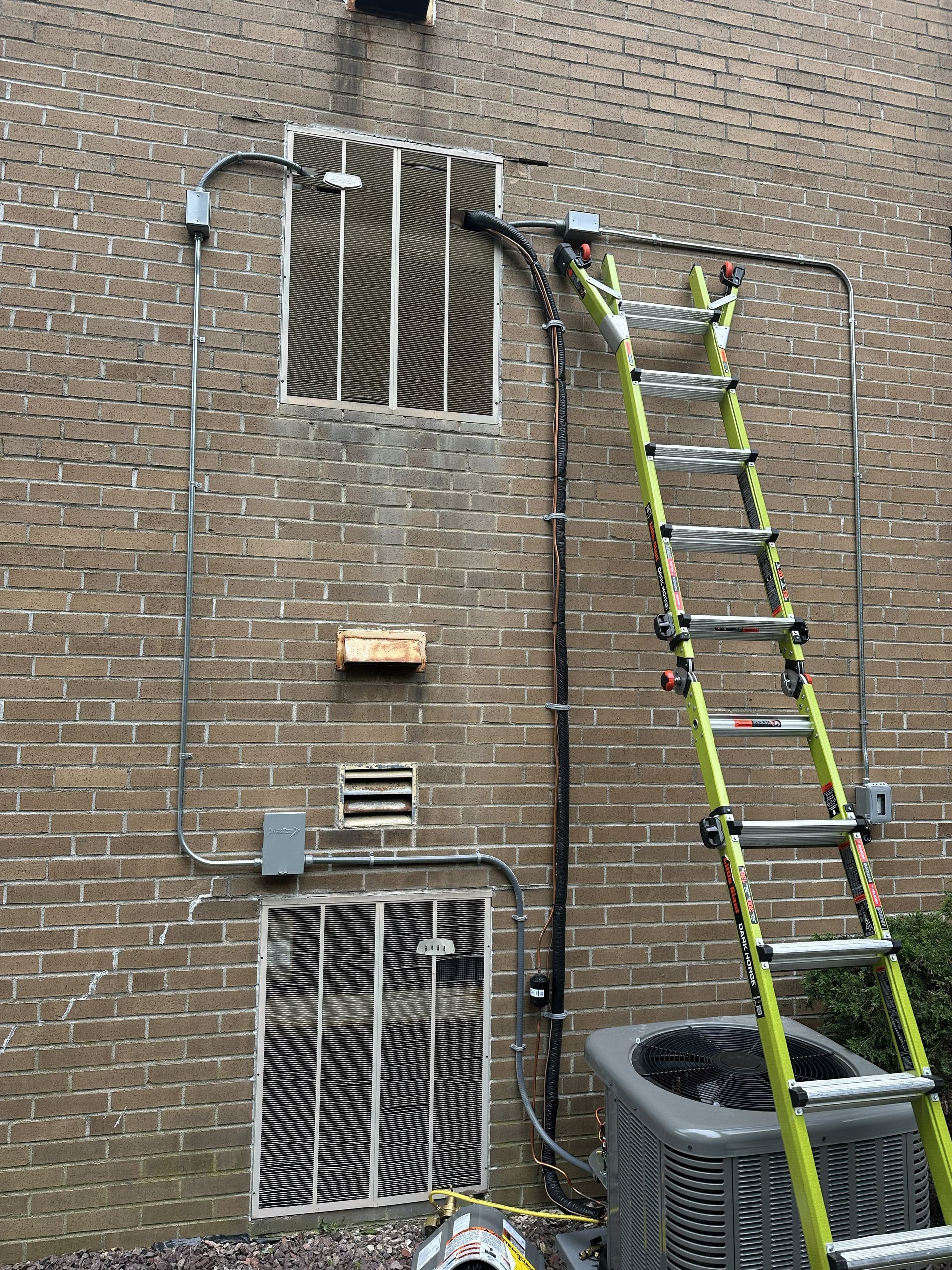 A tall extension ladder leans against a brick wall next to HVAC vents, exposed electrical conduits, and a compressor unit.