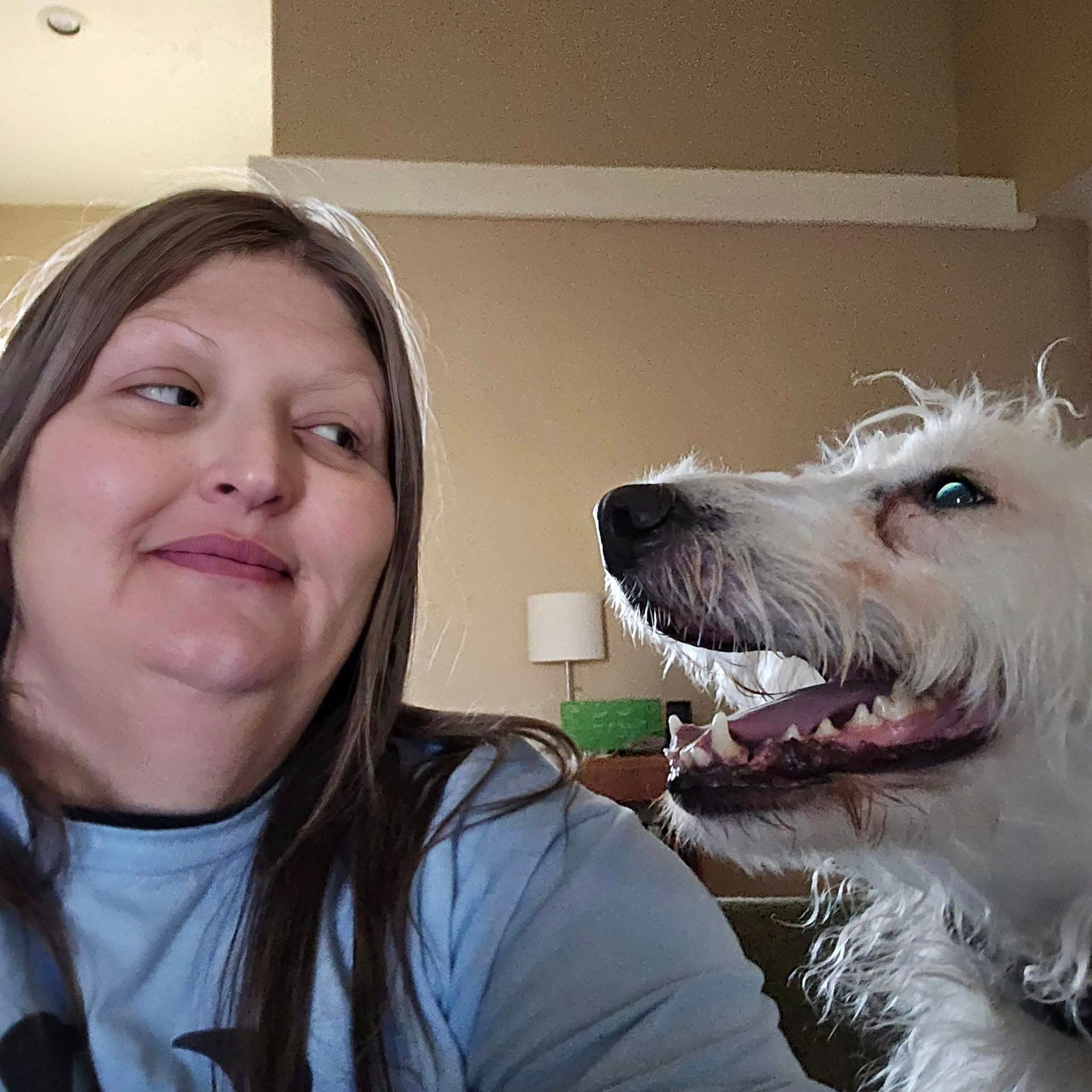 Woman smiles beside a large white dog with its mouth open, indoors.