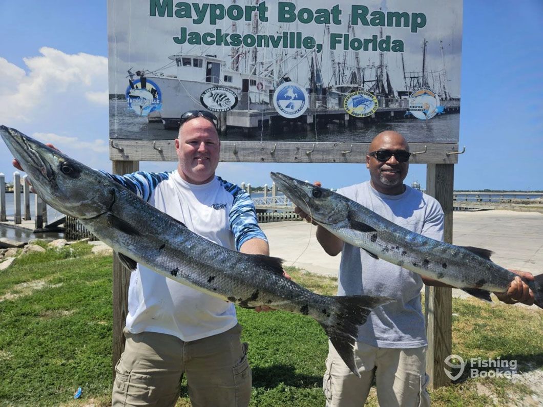 Two men are holding large fish in front of a mayport boat ramp sign.