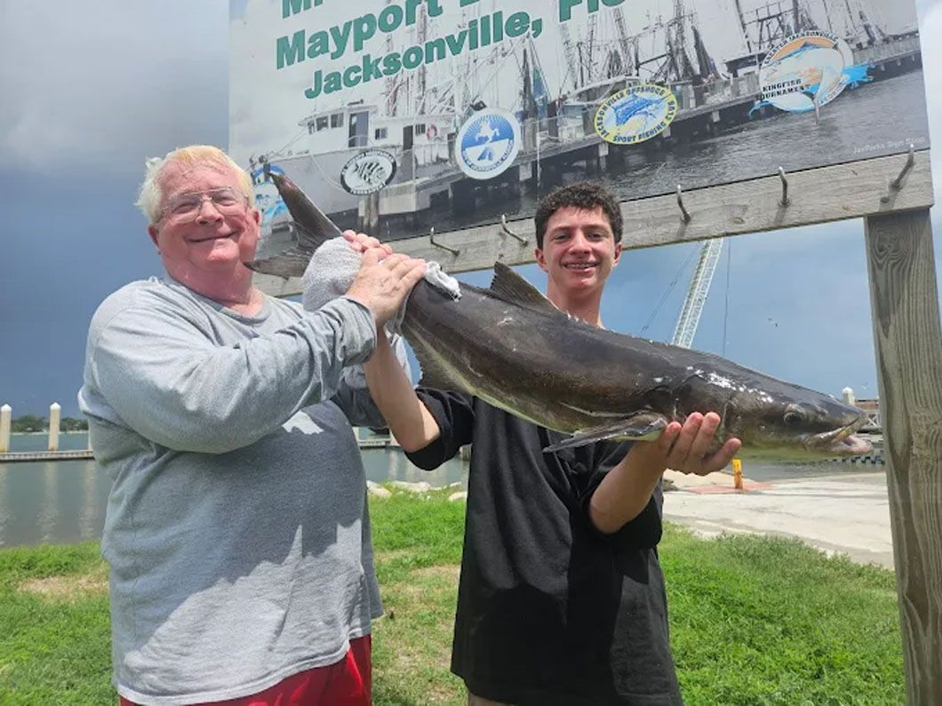 Two men are holding a large fish in front of a sign that says mayport jacksonville