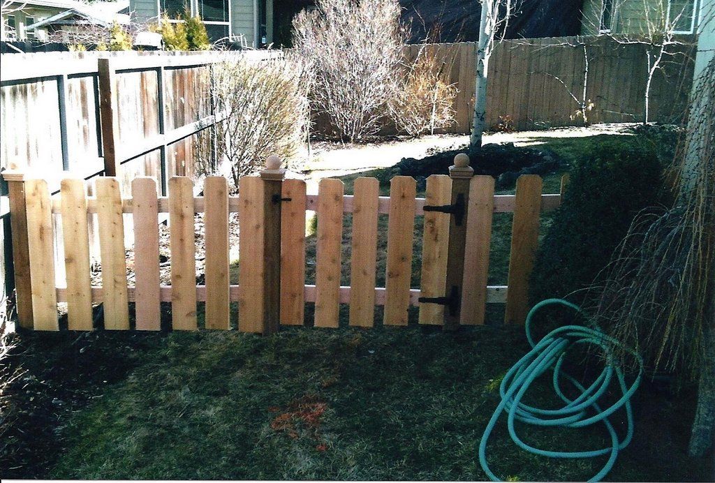 Wooden picket fence with gate in a backyard, brown with black hinges and a green hose nearby.