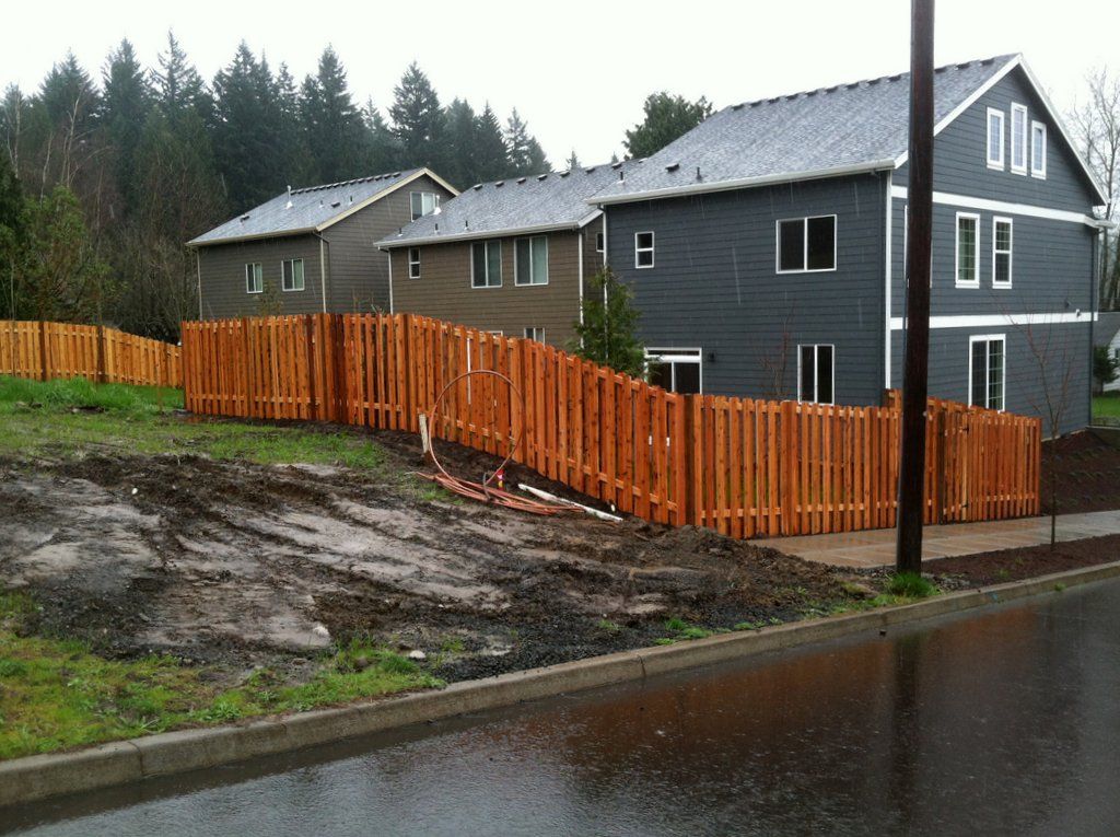 Wooden fence bordering a grassy area with three multi-story houses in the background on a rainy day.