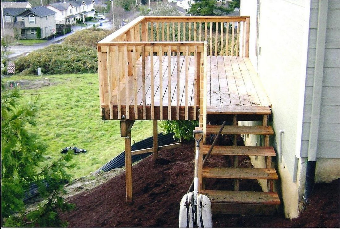 Wooden deck with stairs attached to a house. Brown wood, railings, and steps over a grassy hill.