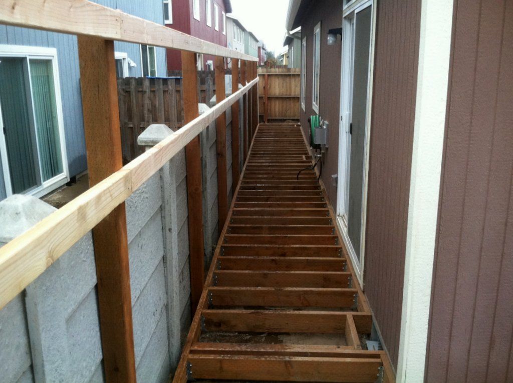 Wooden walkway under construction, between a fence and a building with a sliding glass door.