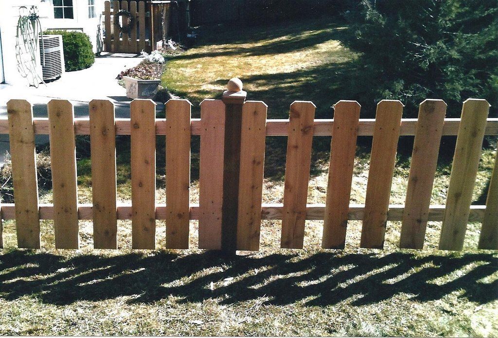 Wooden picket fence with open gate casting shadows on grass in front of a house.