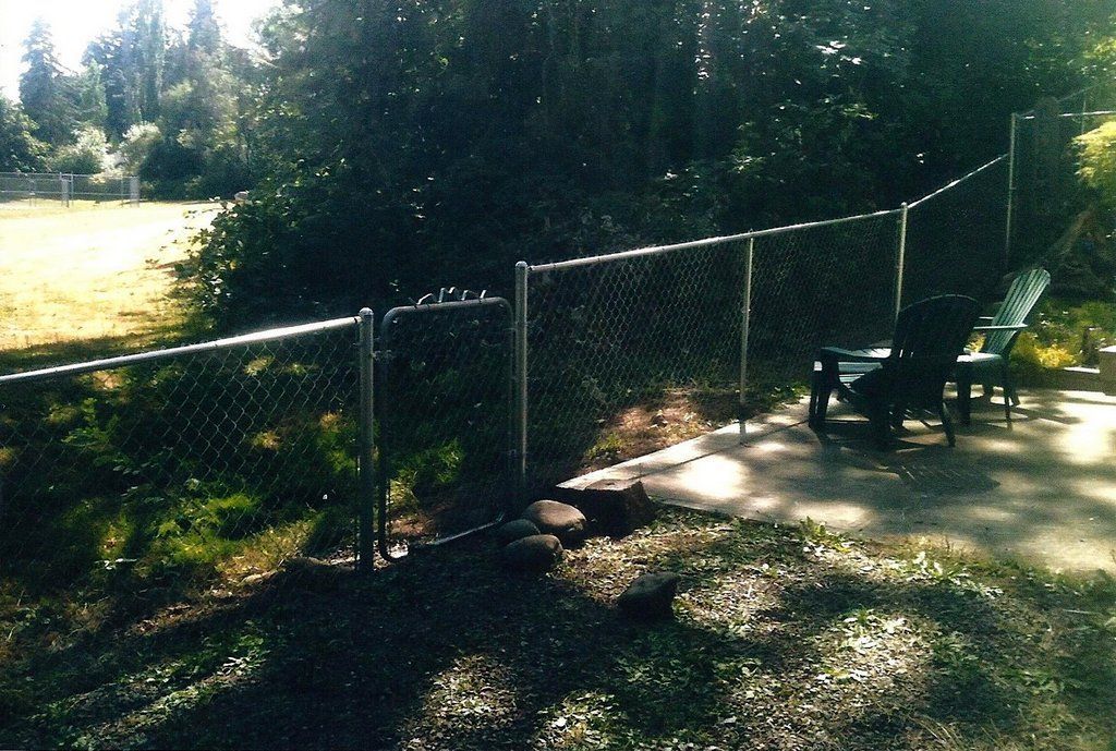 Chain link fence encloses a patio with two lounge chairs, and a grassy area with trees and a field in the background.
