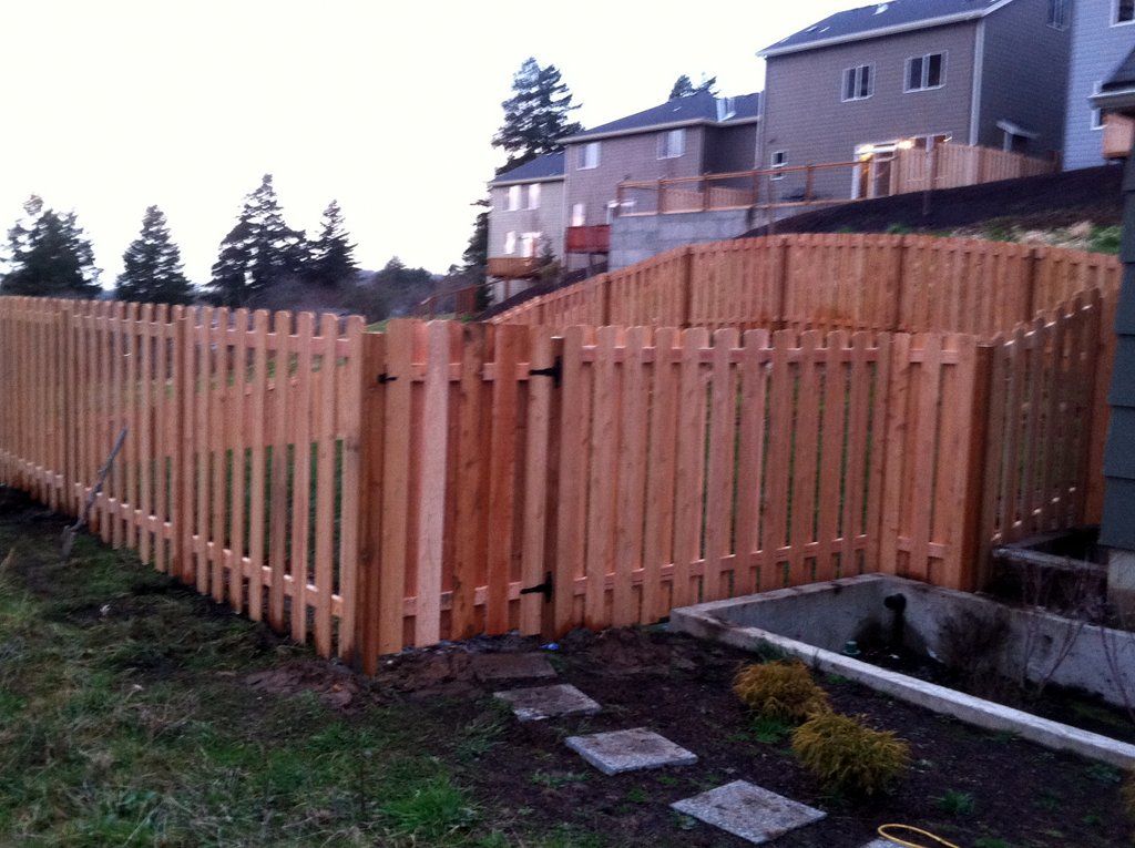 Wooden picket fence with a gate, on a hillside, surrounding a yard, in front of houses.