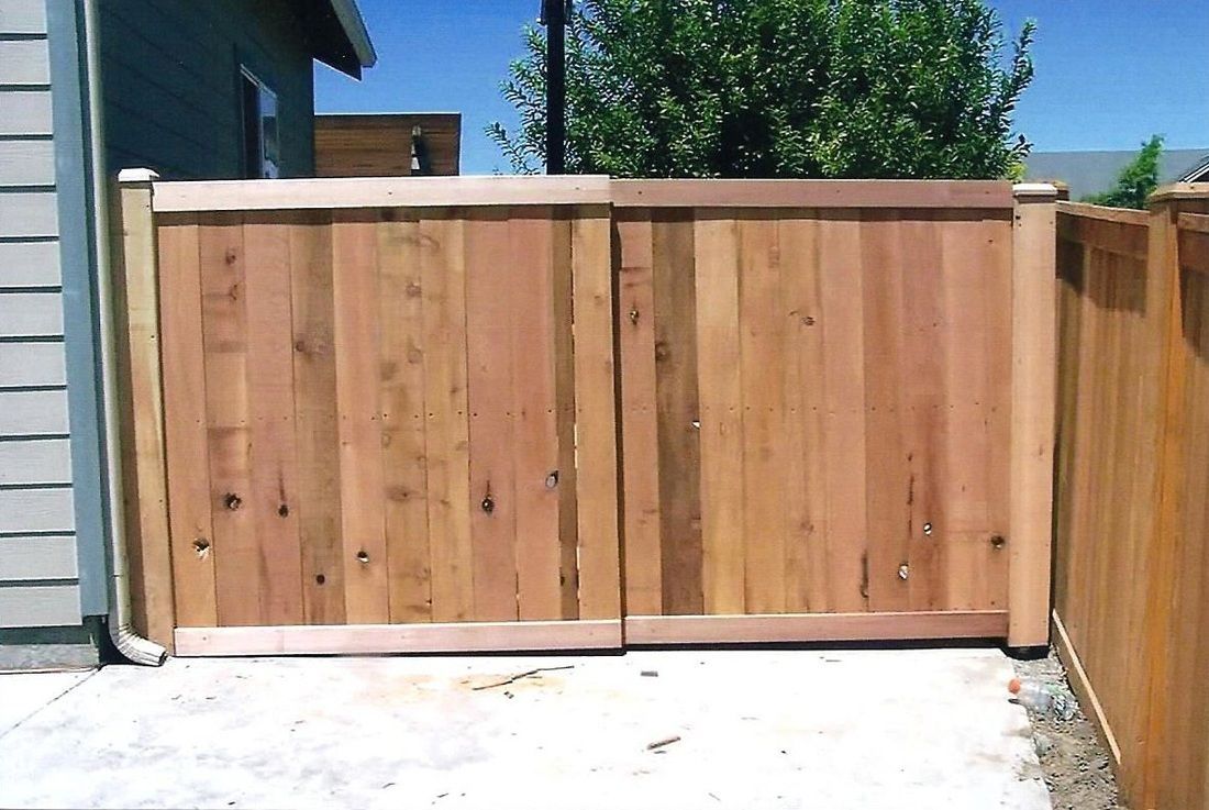 Wooden double gate in a concrete driveway, against a light-colored house and wooden fence.