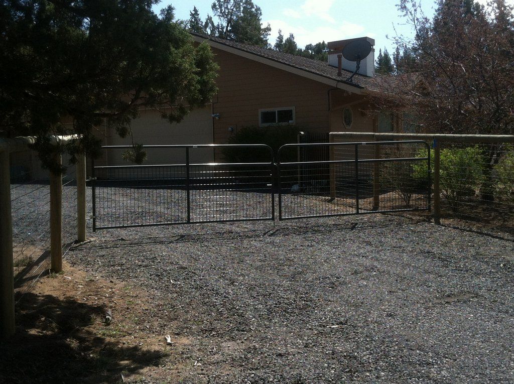 A house with a black metal fence in front on a gravel yard, with trees in the background.