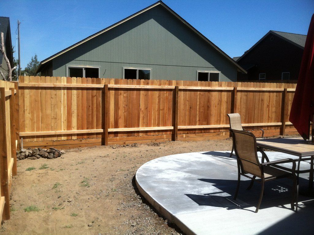 Wooden fence surrounds a backyard patio area with a table and chairs. A house is in the background.