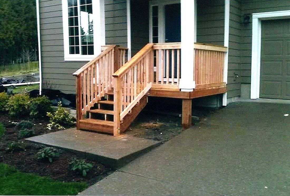 Wooden porch with stairs and railing, concrete path, and house with a green exterior.