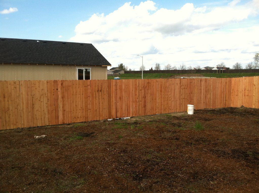 Wooden fence around a house in a yard with sparse vegetation and a cloudy sky.