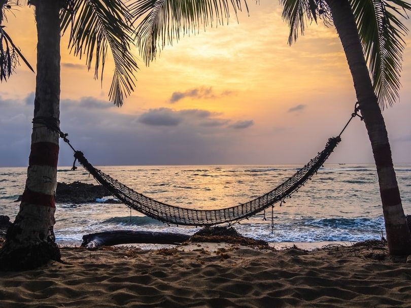 A hammock is hanging between two palm trees on a beach at sunset.