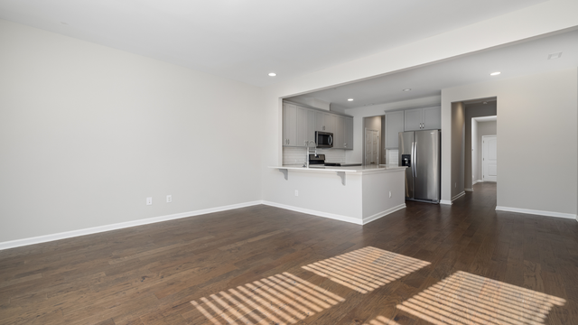 An empty living room with hardwood floors and a kitchen in the background.