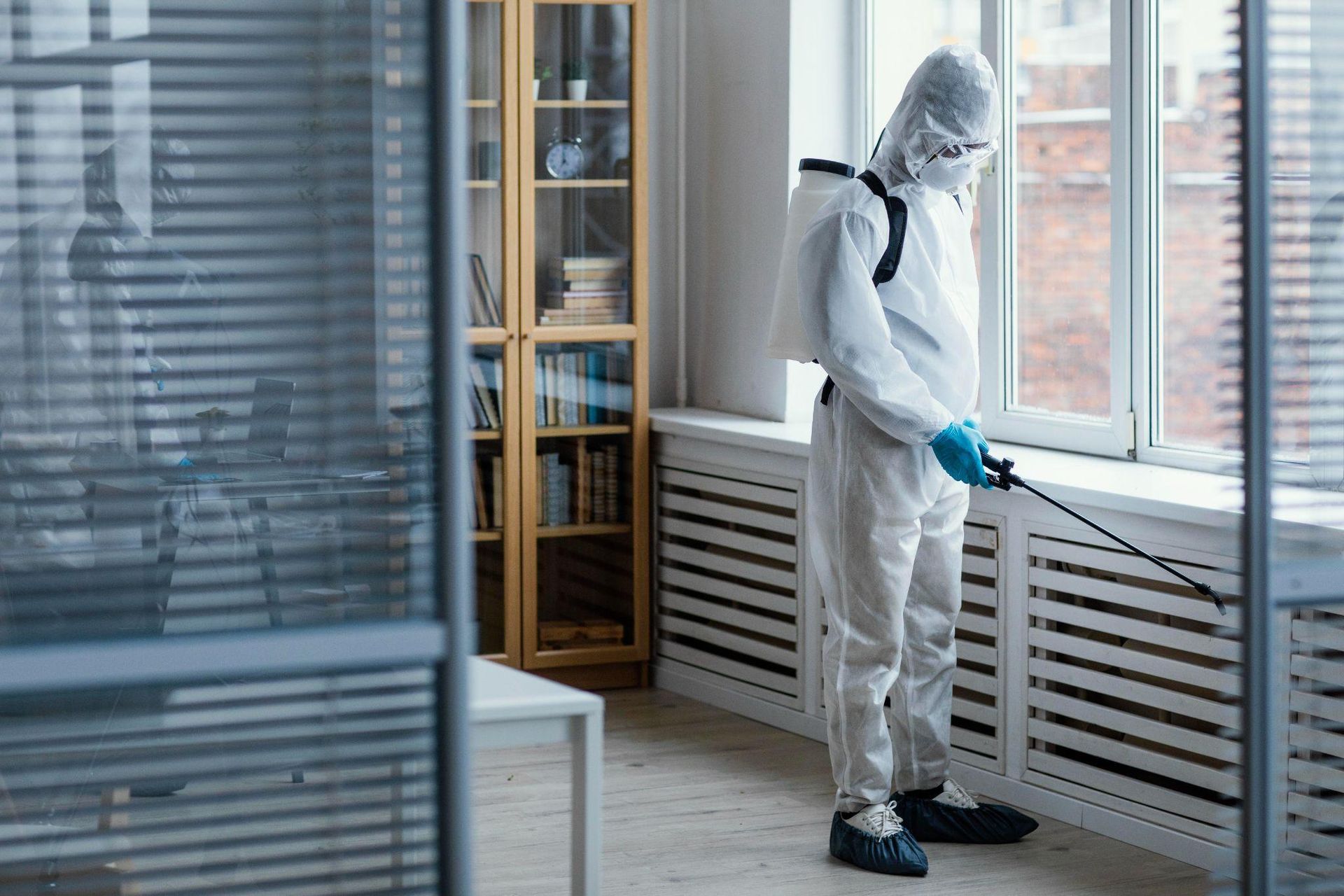 Person in hazmat suit sanitizing a room with a sprayer near a window.