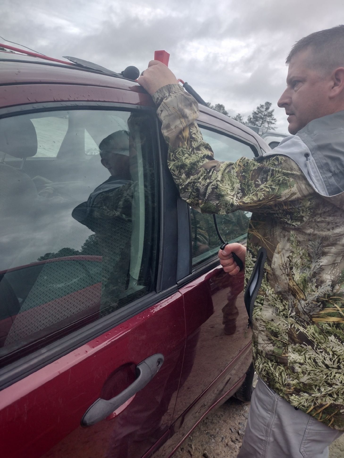 A man in a camo jacket is standing next to a red car.