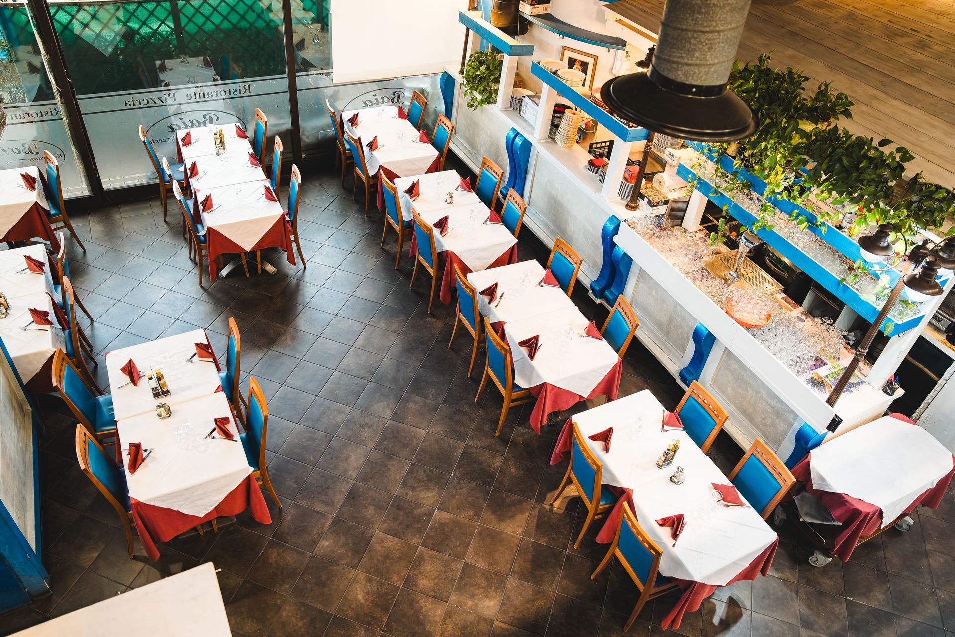 Vista dall'alto di un ristorante con tovaglie bianche, tovaglioli rossi, sedie blu e arredamento in stile greco.
