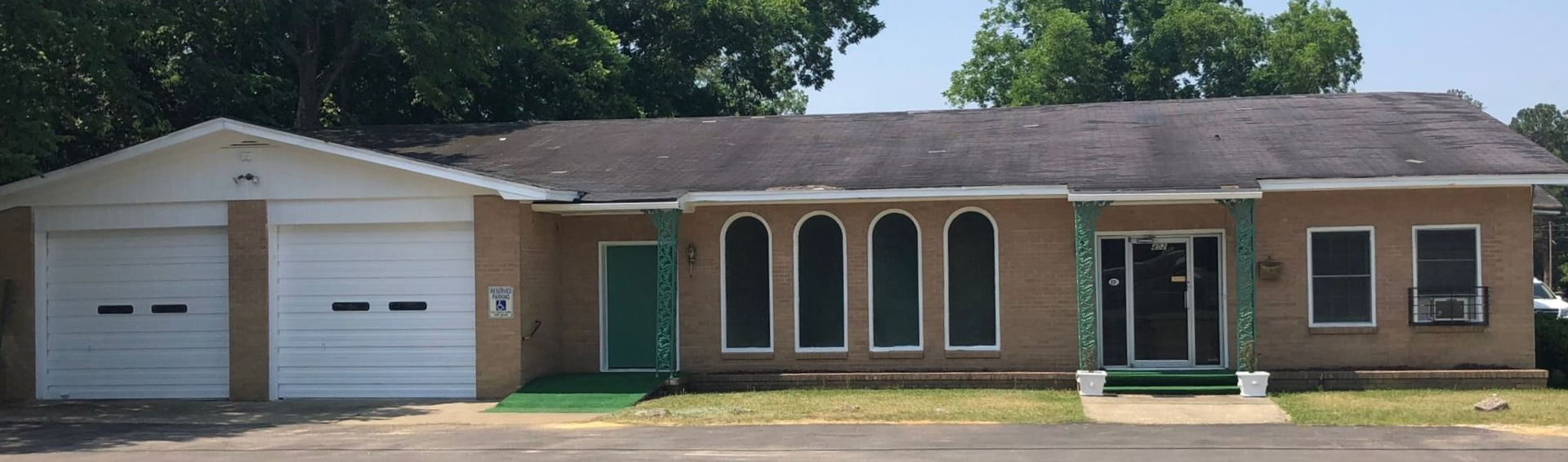 A tan, single-story building with a dark roof has two white garage doors and arched windows. Green door. Trees in the background.