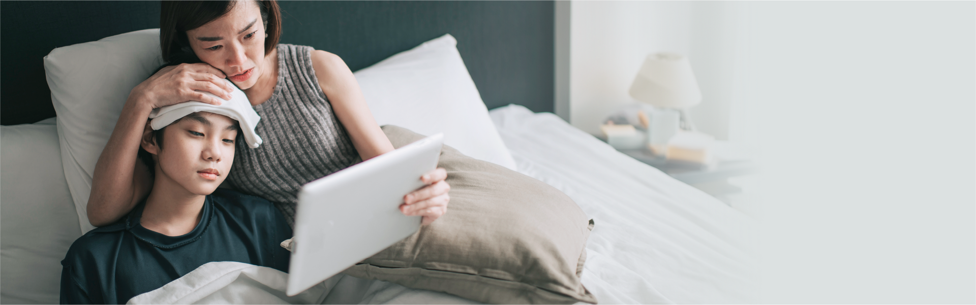 A person rests in bed with a cool compress on their forehead while another person holds a tablet over them.