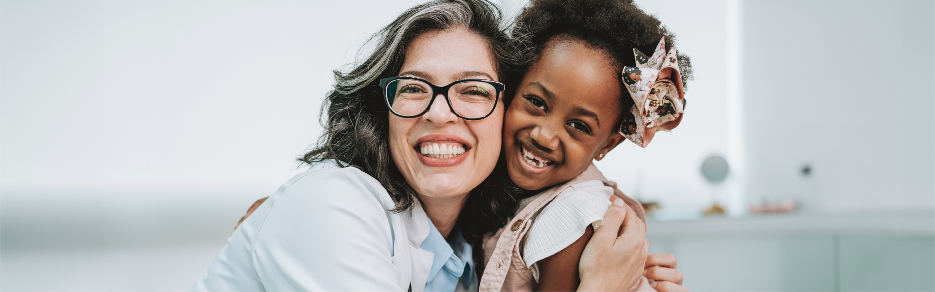 Woman in glasses hugs a young child. They are smiling in a brightly lit room.