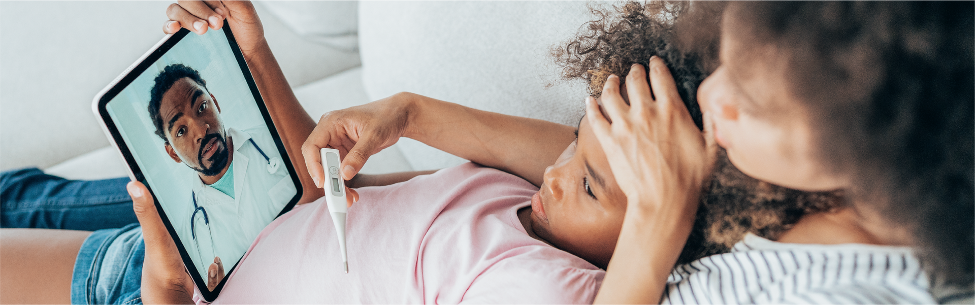 A sick child consults with a doctor via telehealth, as a parent checks the child's forehead with a thermometer.
