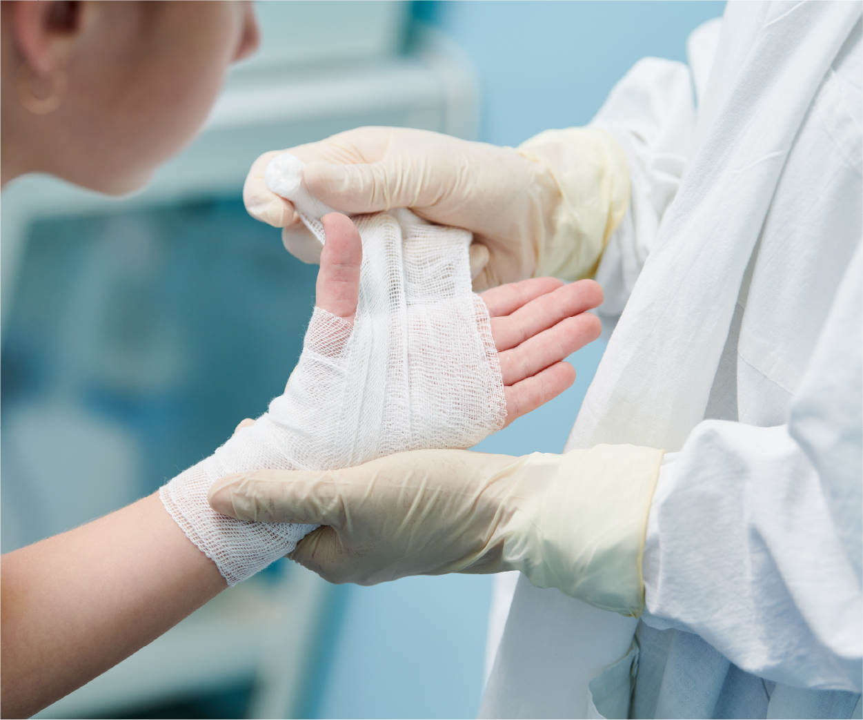 A gloved hand wrapping a gauze bandage around a child's injured hand in a medical setting.