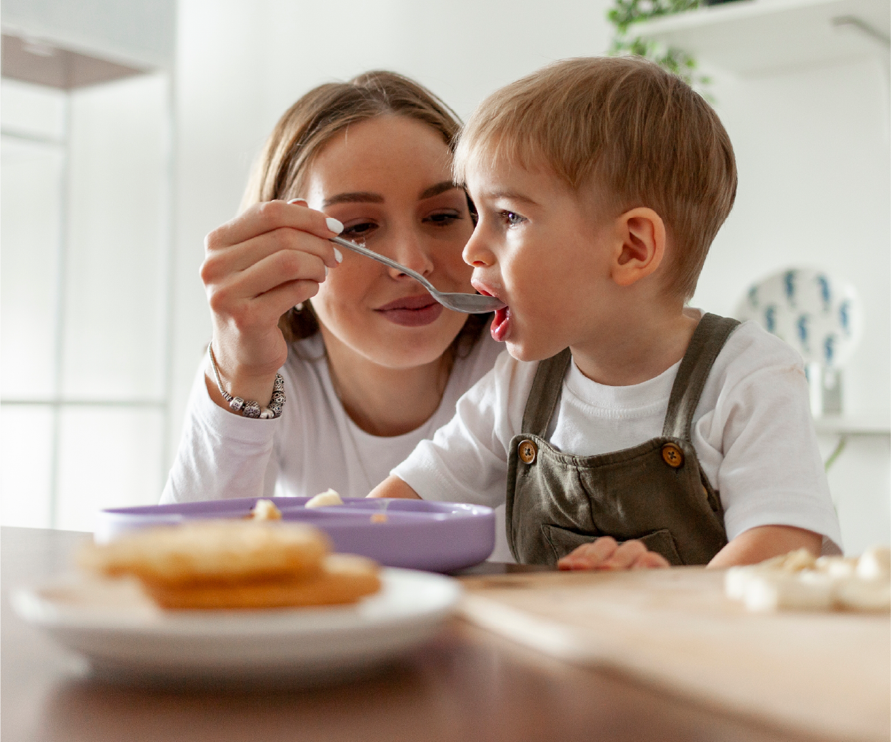 Woman feeding a child with a spoon, sitting at a table with food.