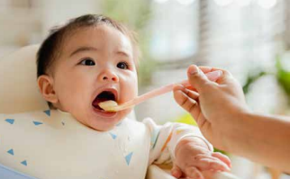 Baby being fed with a spoon, wearing a white bib and looking up with mouth open.