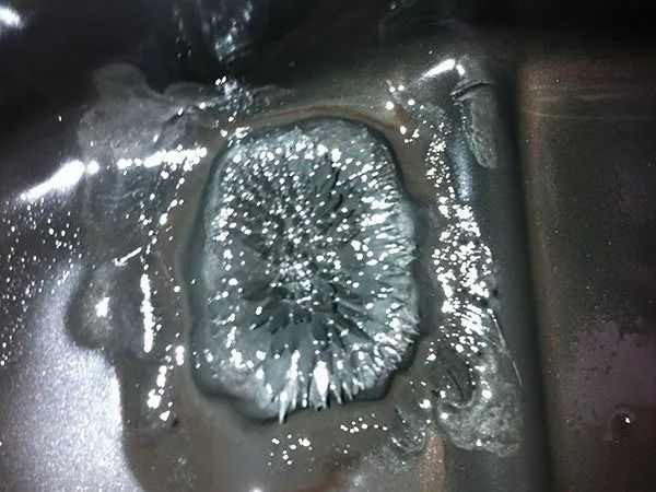 Clumped metal shavings forming a spiky, oval shape in a metallic surface; surrounded by liquid.