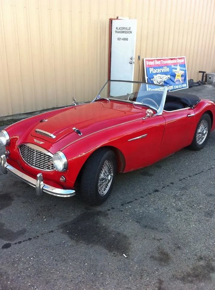 Red classic convertible car parked in front of a building with a sign and open door.