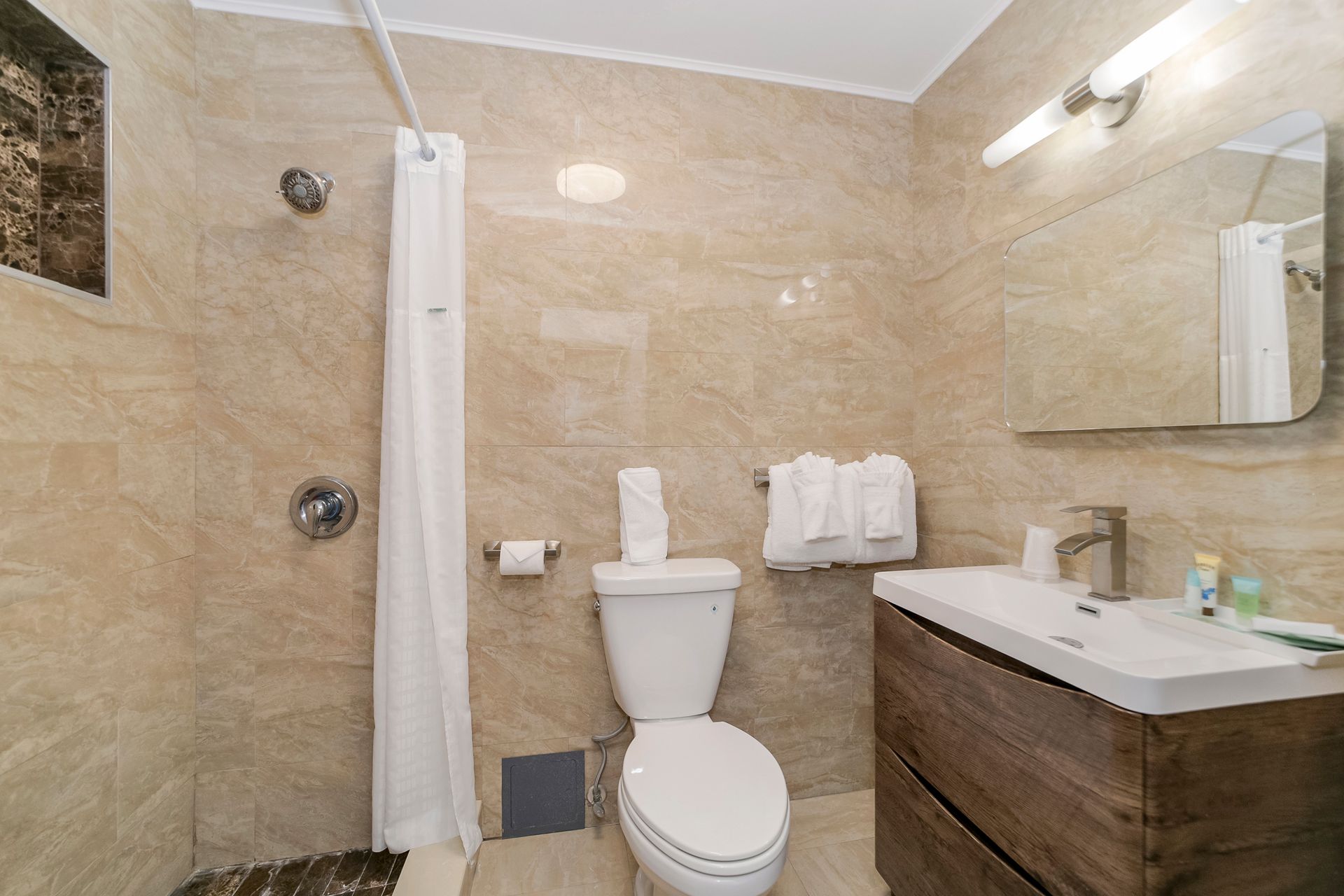 Bathroom with beige tile walls, shower, toilet, sink with wooden cabinet, and mirror.