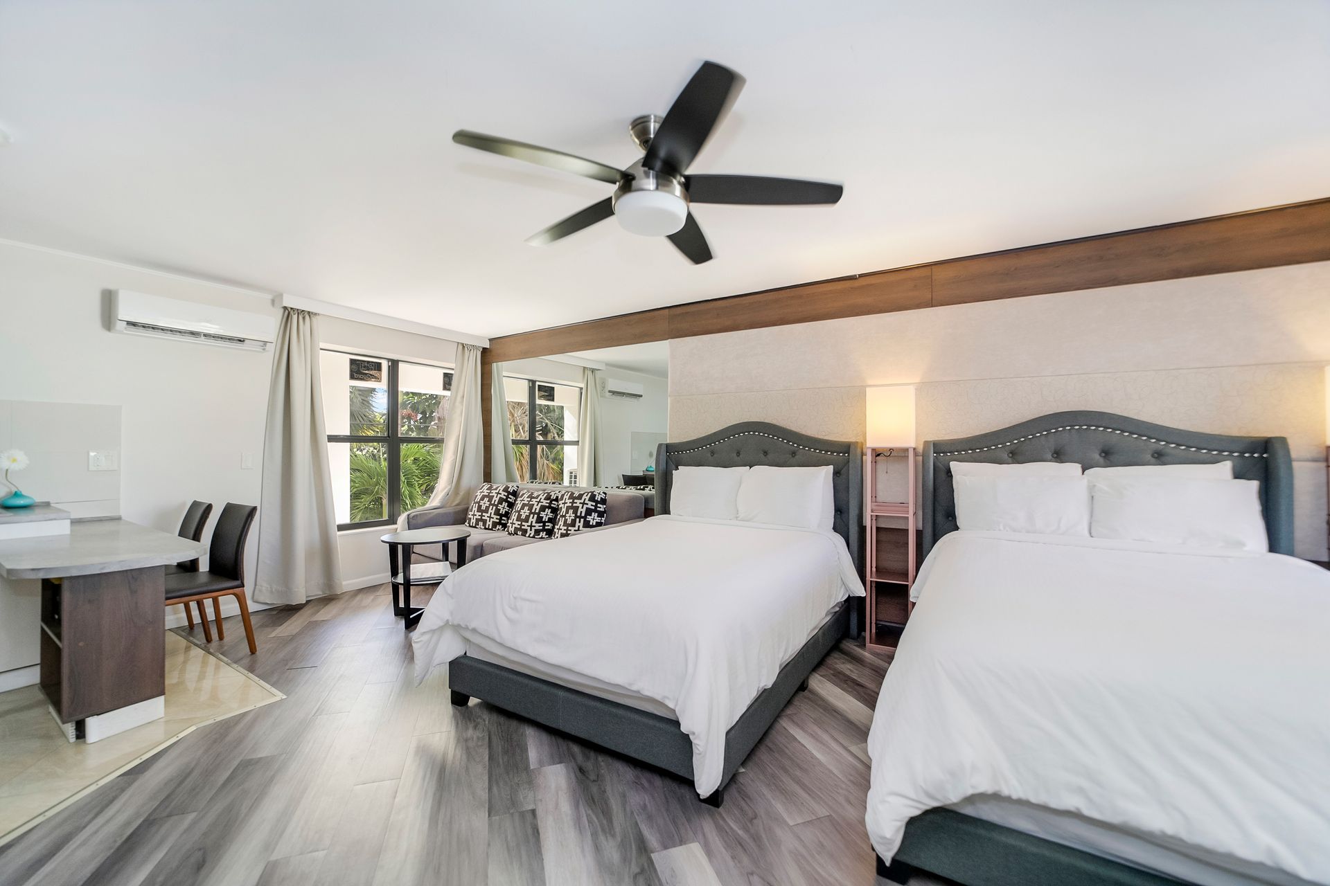 Hotel room with two beds, small sitting area, and a table. Light wood floor, white walls.