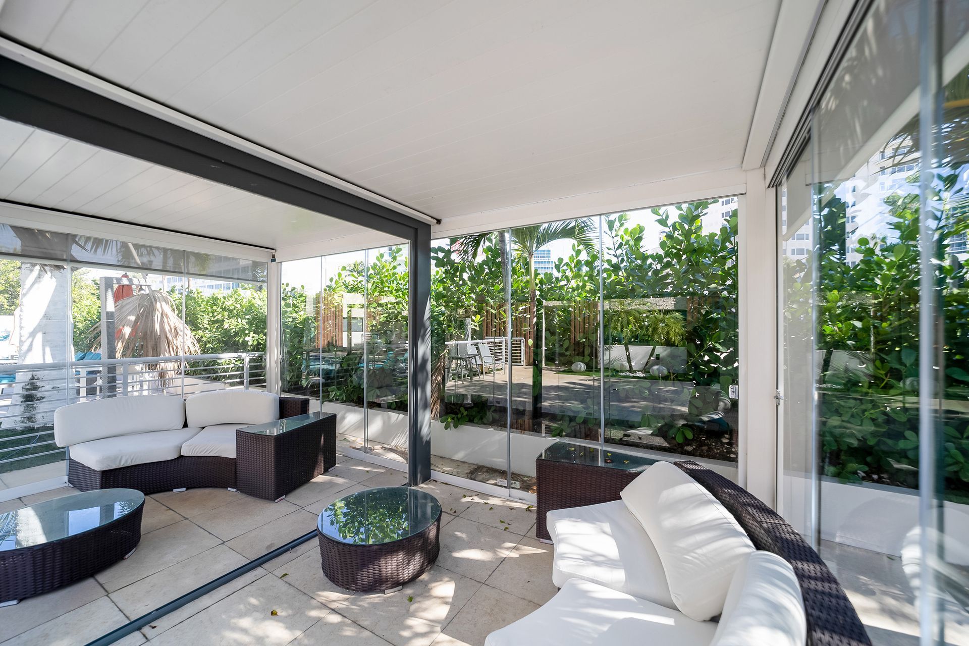 Patio with white sofas, glass walls, and a view of greenery.