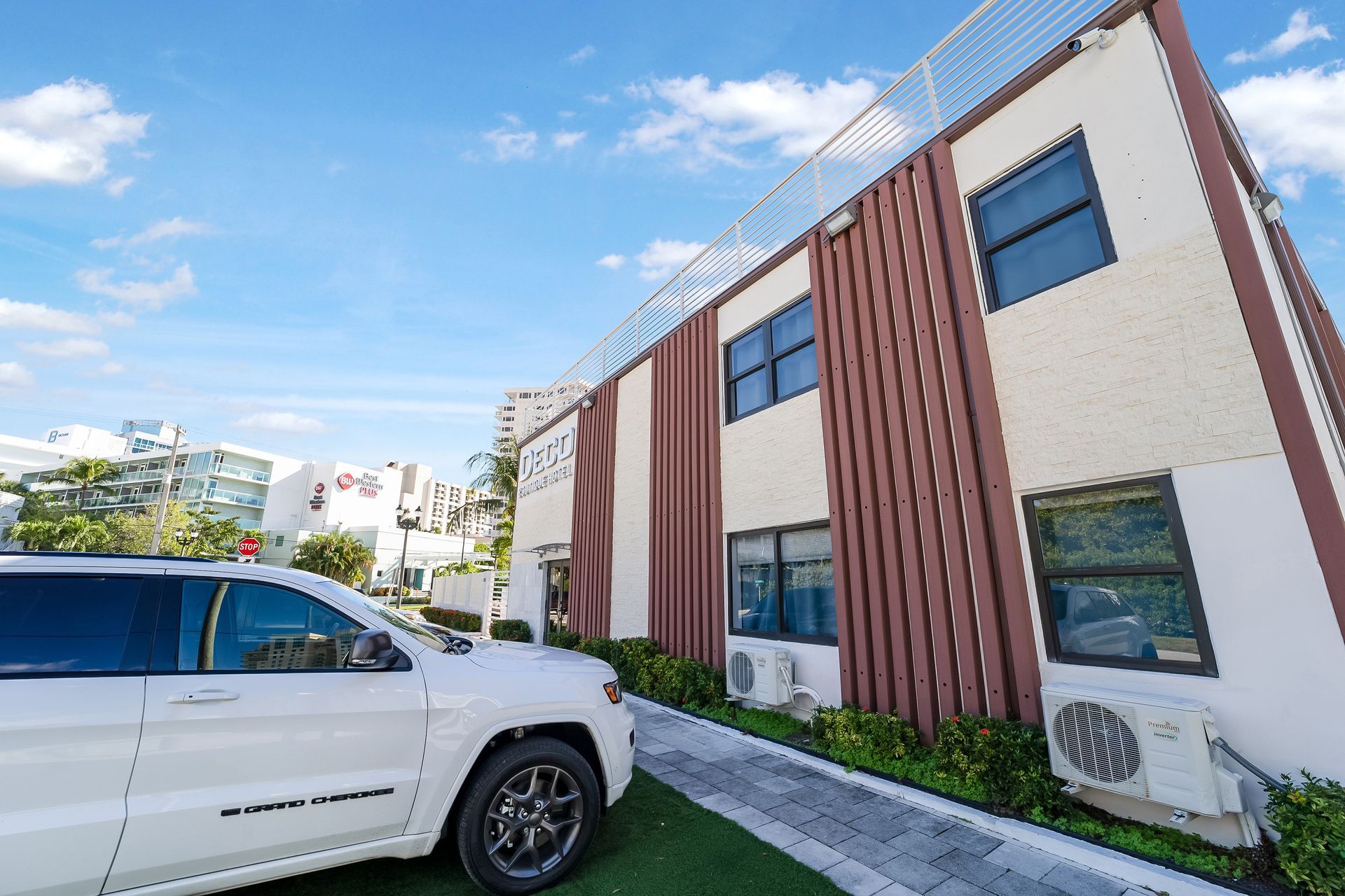 White SUV parked beside a two-story building with brown vertical panels, green grass, and blue sky.