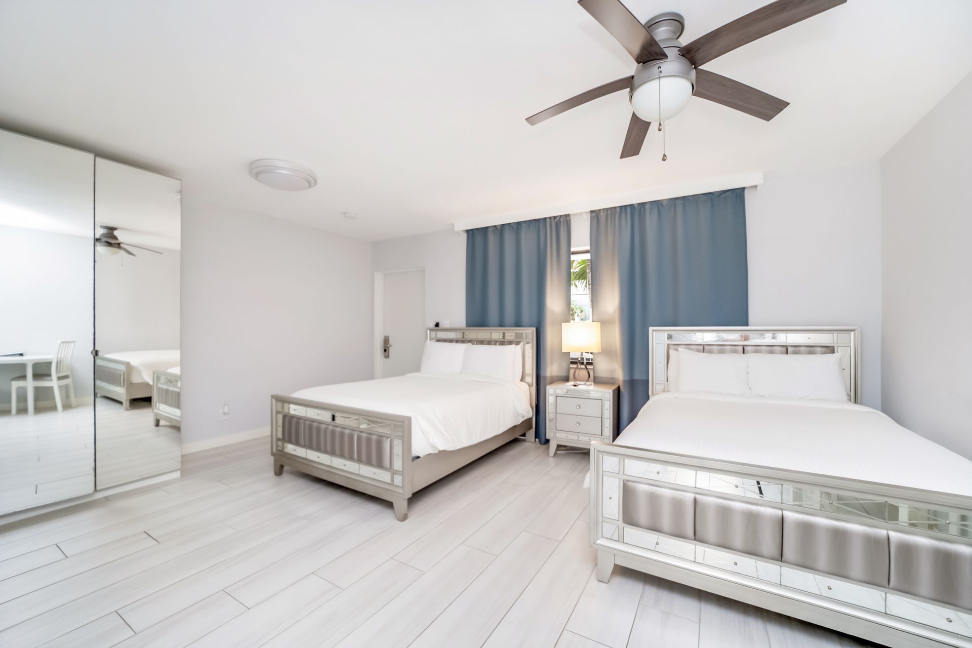 Bedroom with two beds, large mirror, blue curtains, ceiling fan, and white walls and flooring.
