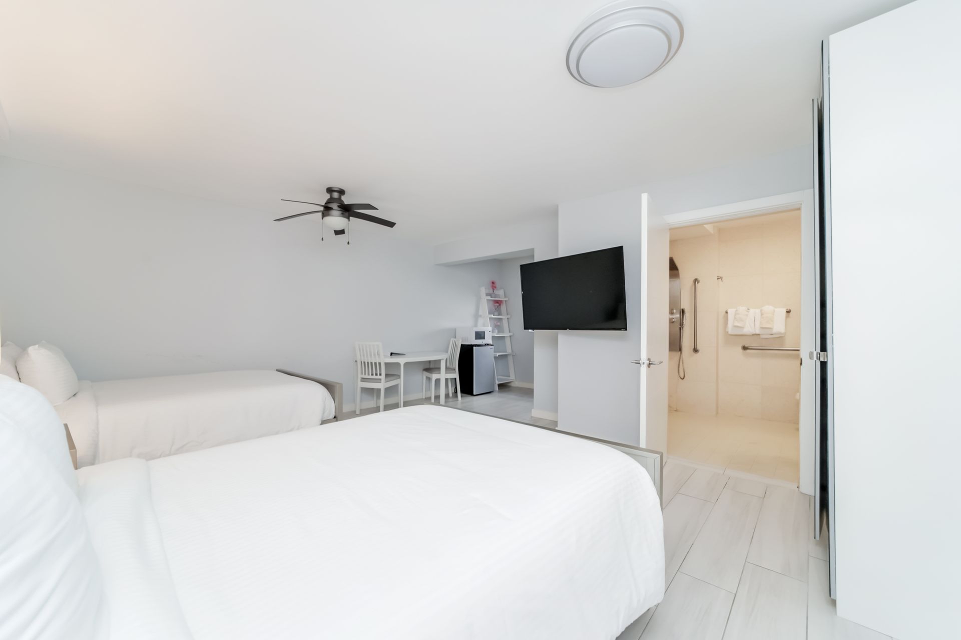 A hotel room with two beds, a small table, a TV, and a door to a bathroom; all in white.
