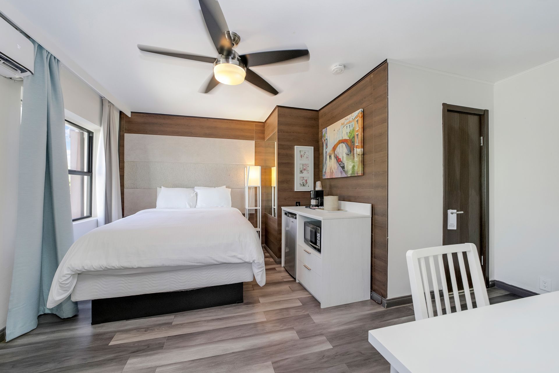 A small hotel room with a bed, kitchenette, and table. Dark wood accents and a ceiling fan are visible.