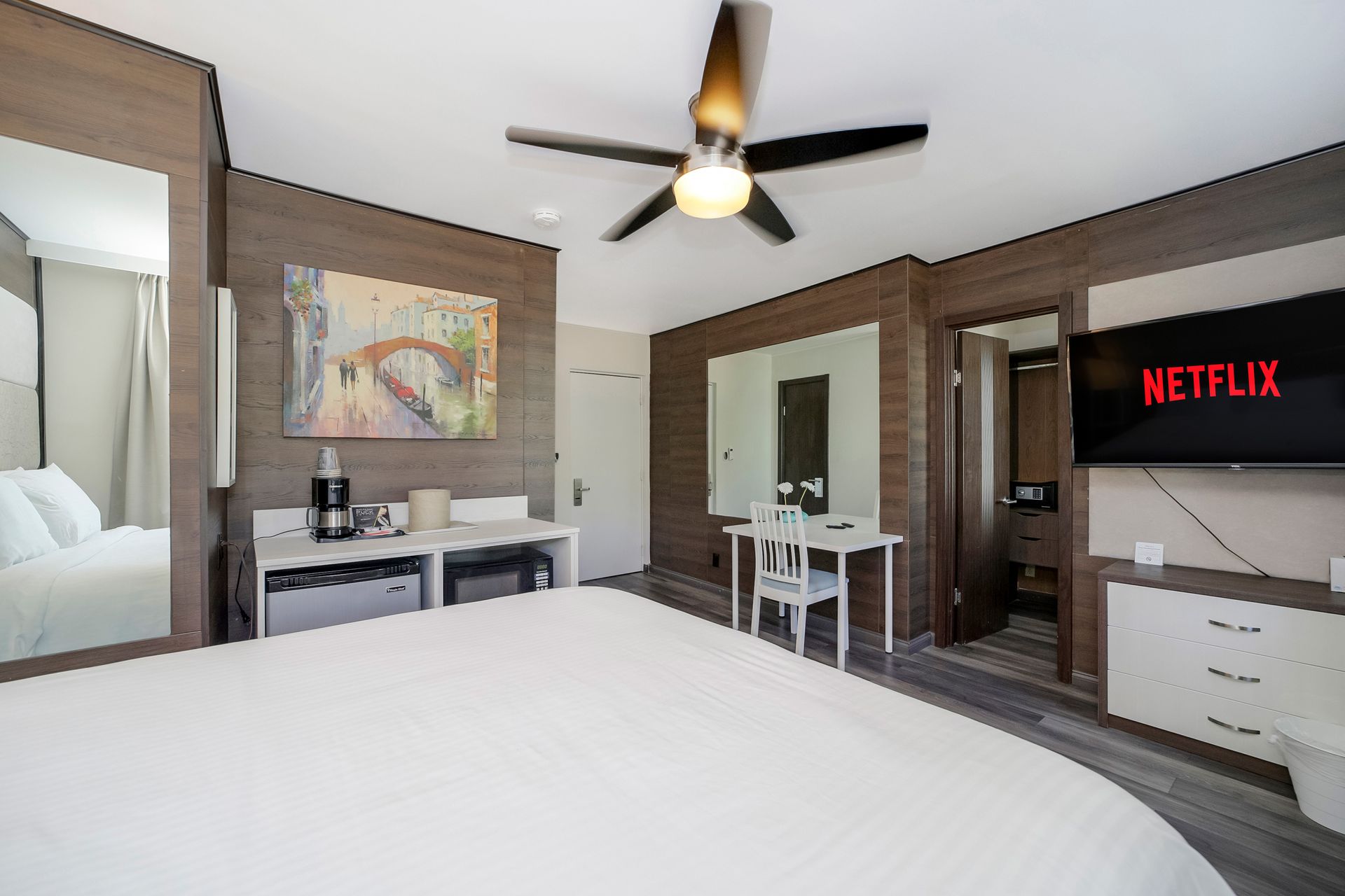 Hotel room interior with bed, TV, desk, artwork, and bathroom door. Brown and white decor.