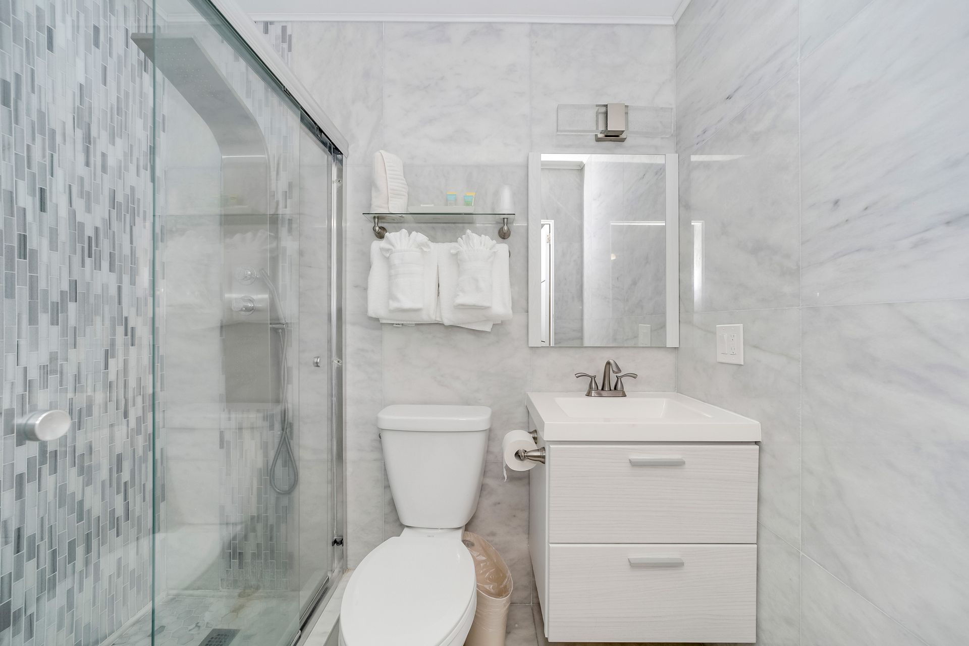 Small, white-tiled bathroom with a glass shower, toilet, sink, mirror, and towel rack.