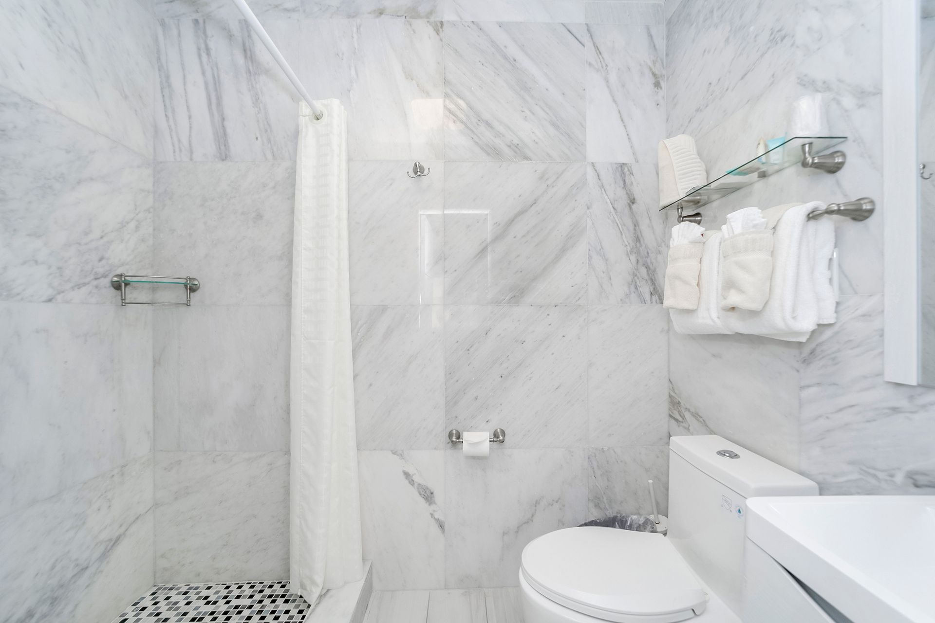 White marble bathroom with shower, toilet, and vanity. White towels on rack.