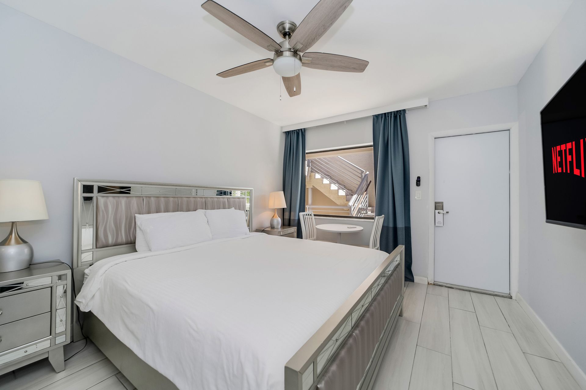 Bedroom with a bed, nightstands, TV, and blue curtains. Ceiling fan and a window are visible.