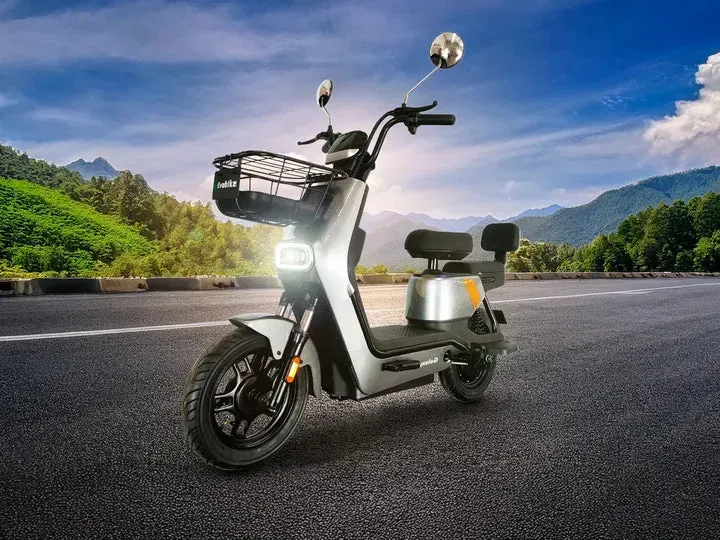 Patinete eléctrico plateado con cesta en una carretera, montañas al fondo.