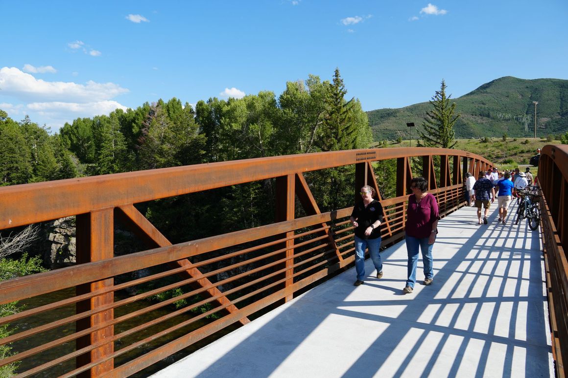 People walk across a rust-colored metal truss bridge over a wooded area on a sunny day in the roaring fork valley