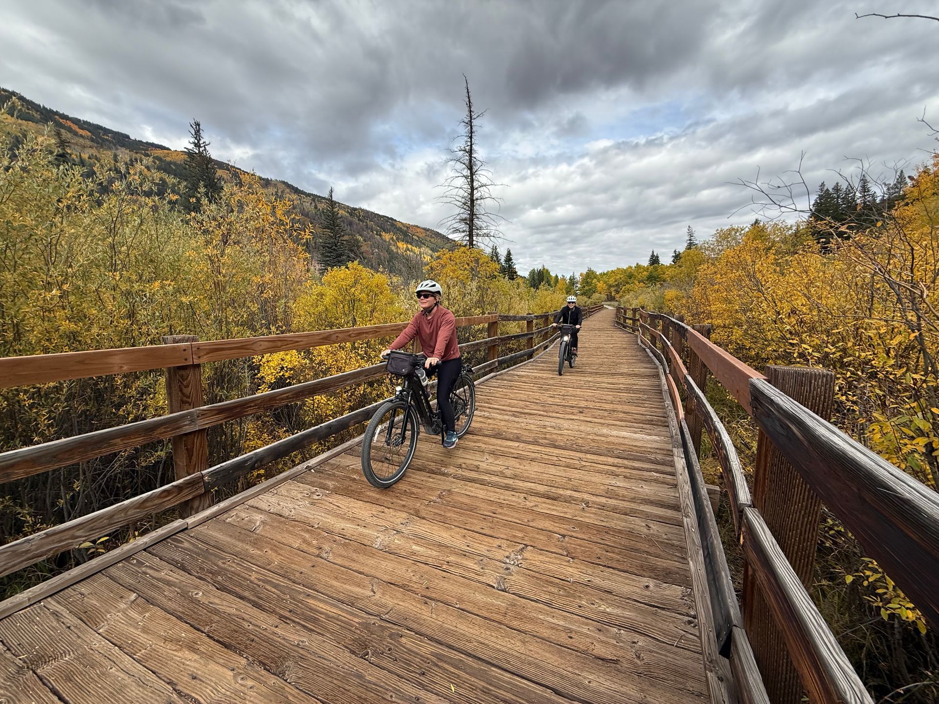 Cyclists ride across a wooden boardwalk through autumn trees under a cloudy sky  with slow & say hello in colorado